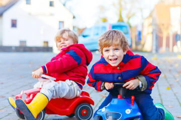 Two little kids boys in colorful clothes and rain boots driving toy cars. Siblings making competition, outdoors. Active leisure for children on autumn day.