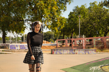 Cute young girl in punk style with white headphones and inline skates skating looking down.