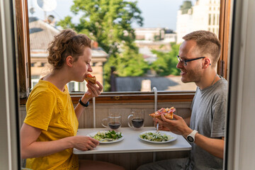A young happy couple is having meal, dinner, enjoying pizza, salad and drinking Kvass at home on...
