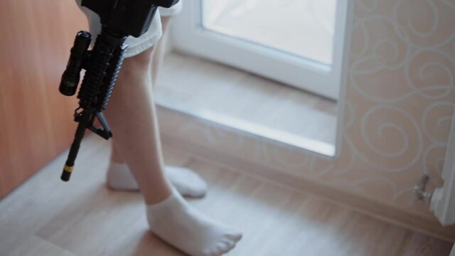 Unrecognizable caucasian man in socks and shorts with hairy legs holds assault rifle. Terrorist stands near balcony door and then walks across room. Barefoot woman in dress standing in apartment.