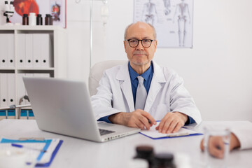 Portrait of senior man specialist physician looking into camera sitting at desk in meeting room working at medical expertise. Doctor prescribing pill medication treatment writing on clipboard
