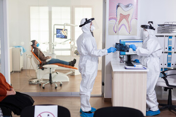 Dental receptionist dressed in coverall face shiled giving doctor patient x-ray keeping social...