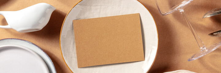 Fototapeta premium Invitaion mockup panorama, top shot with tableware and glasses. RSVP card on a plate, with copy space. Special occasion design template