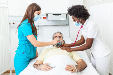 Obraz premium Nurse puts oxygen mask on elderly man patient lying in the hospital room bed while African American doctor examining patient with stethoscope, coronavirus covid 19 protection concept
