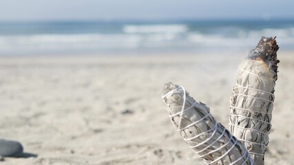 Dried white sage smoke, dry smudge stick burning, soft focus bokeh. Aroma smudging close up on ocean beach, aromatherapy meditation by sea water waves on sandy coast. Yoga, peace, harmony and ayurveda