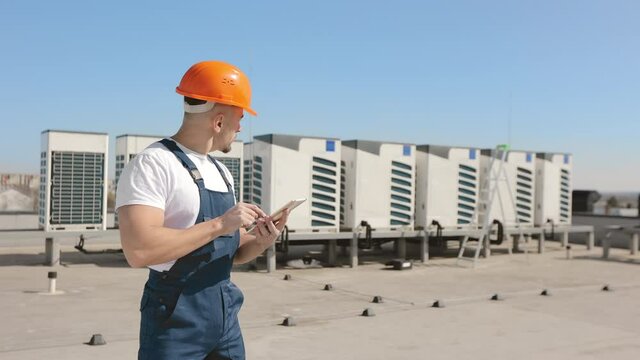 Portrait Of A Serious Young Engineer Is Working On The Tablet And Looking At The Air Conditioning Systems. He's On The Roof Of A Business Center. He Is Wearing Work Clothes And A Hard Hat. Nice Sunny