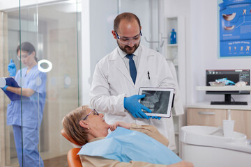 Fototapeta premium Stomatolog in uniform during consultation of senior woman explaining diagnostic. Medical teeth care taker holding patient radiography on tablet pc near patient standing up.