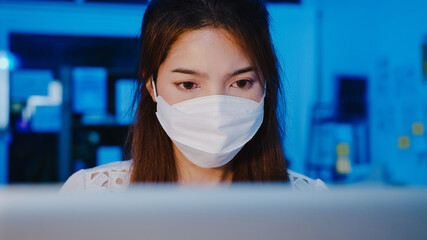 Happy Asia businesswoman wearing medical face mask for social distancing in new normal situation for virus prevention while using laptop back at work in office night. Life and work after coronavirus.