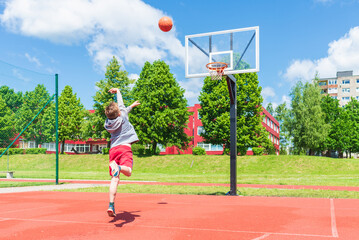 Cheerful Boy performs shot to the basket at basketball game on the playground during sunny summer...