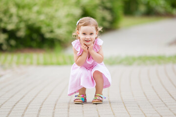 Happy little girl in pink dress squatting on pavement at city park in warm summer day. Looking at...