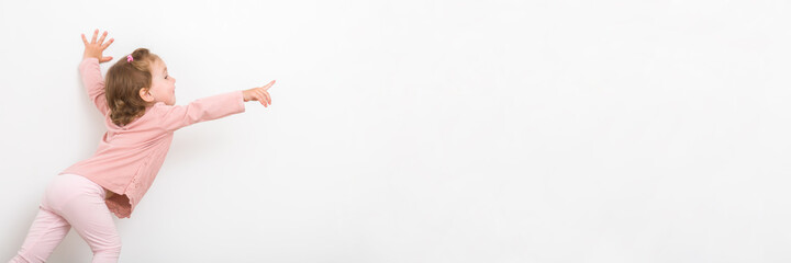 Happy little girl in pink clothes stretching and pointing with finger to gray wall background. 2 years old toddler. Side view. Wide banner. Empty place for text, quote or saying.