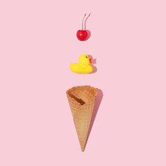 Creative composition with ice cream cone, yellow rubber duck and bright red cherry on pastel pink background. Summer dessert art direction. Summer food minimal concept.