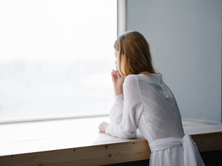 red-haired Woman in a white dress enriched herself on the windowsill near the window