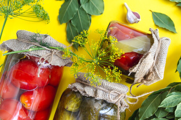 Glass jars with homemade pickled tomatoes, sealed with metal lid on yellow background. pickled cucumbers, dill sprigs and bay leaves