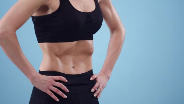 Athletic woman with abs puts her hands on her waist and draws in stomach on blue background, front view, closeup. Yoga concept. Exercise vacuum