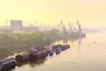 A foggy morning on the Ob River in Novosibirsk. Berths and ships on the calm surface of the water, the shore of the city with port cranes in the haze on the horizon. Siberia, Russia