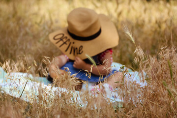A little infant girl in a big straw hat with a word Offline written on it sits on a plaid among yellow sunburned grass in a field. Kid having fun in nature in a summer. Baby in field. Family vacation.