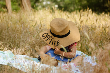 A little infant girl in a big straw hat with a word Offline written on it sits on a plaid among yellow sunburned grass in a field. Kid having fun in nature in a summer. Baby in field. Family vacation. - obrazy, fototapety, plakaty