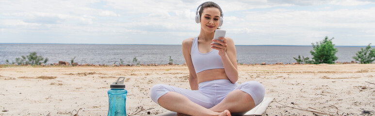 smiling woman in headphones listening music while using smartphone and sitting with crossed legs on yoga mat, banner