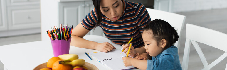 Asian mother and kid drawing near fruits in kitchen, banner
