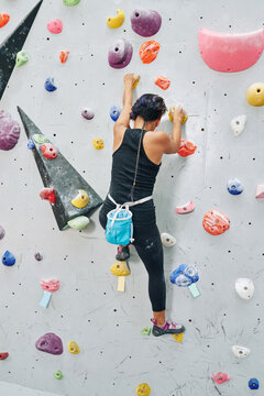 Fit strong young woman climbing up challenging route in rock-climbing gym, view from the back