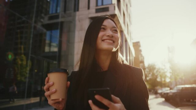 Smiling happy woman walking while using phone hold drink coffee in urban city buildings look around at sunlight. Portrait, cellphone, street, beautiful, travel, mobile, stylish. Slow motion