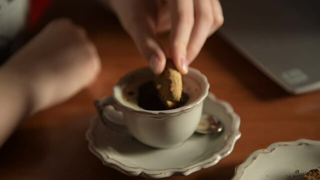 4k close up of female hand Hand dip cookie in cup of aromatic coffee. Vintage coffe cup is placed on a wooden table near laptop, concept of coffee break and slow breakfast, soft focus