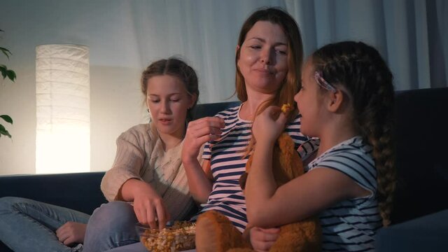 Happy family watching TV at home. Mom and kids are watching tv with popcorn. Happy family watching movie at home. Mom and children are eating popcorn on the couch. Happy family watching movie