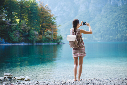 A girl-blogger pictures the landscape on a smartphone. A white Caucasian girl in a pink dress and a stylish backpack on her shoulder makes selfie on the background of a lake with turquoise water