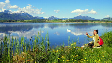 Fototapeta na wymiar junge Frau genießt die stille Natur am Hopfensee im Allgäu am grünen Ufer sitzend bei blauem Himmel 