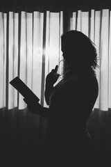 Side view of silhouette woman standing by window reading a book