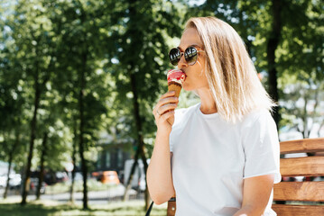Attractive young woman with glasses resting on a park bench and eating ice cream in a waffle cone....