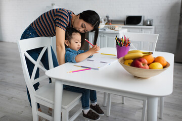 Asian mother kissing head of daughter drawing near fruits at home