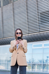 Woman wearing trendy sunglasses walks down the central city street and uses her phone. Pretty summer woman in white jacket walks down the street looking at her mobile phone.