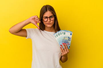 Young caucasian woman holding bills isolated on yellow background showing a dislike gesture, thumbs down. Disagreement concept.