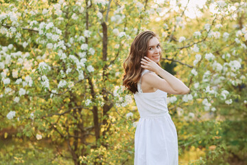 Fototapeta na wymiar A close-up portrait of a happy pretty gentle smiling young woman with a hairstyle in a white cotton dress having fun walking alone enjoying the smell of blooming flowers in a summer green outdoor park