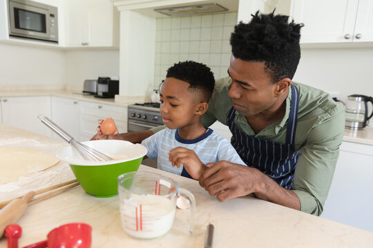 African american father and son baking in kitchen breaking eggs into mixing bowl