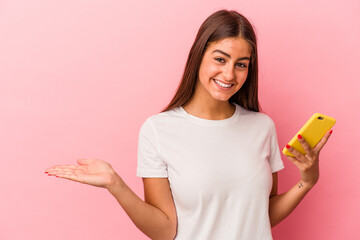 Young caucasian woman holding a mobile phone isolated on pink background showing a copy space on a palm and holding another hand on waist.