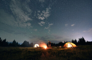 Illuminated camp tents and couple near campfire. Young woman and man travelers standing near bonfire under magical blue sky with stars. Concept of travelling, night camping and relationship.