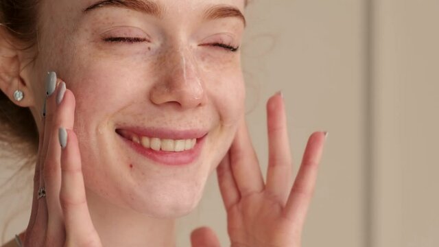 Close up video of a freckled caucasian woman massaging her face with a cream smiling