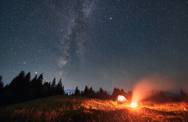 Fototapeta na wymiar Wide angle view on beautiful landscape in the mountains. Tourist tent and campfire under night starry sky with Milky way. Mountains range behind spruces. Concept of night camping and astrophotography