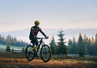 Happy cyclist in cycling suit riding bike on mountain road with coniferous trees and hills on background. Man bicyclist wearing safety helmet and glasses while enjoying bicycle ride in mountains.