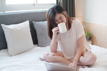 Smiling Asian businesswoman is drinking a cup of coffee and working with laptop in bedroom on holiday. She is online working from home with technology during the Covod-19 outbreak concept.