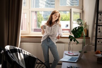 Young beautiful brunette woman drinking morning coffee while standing by the window in the apartment near table with laptop