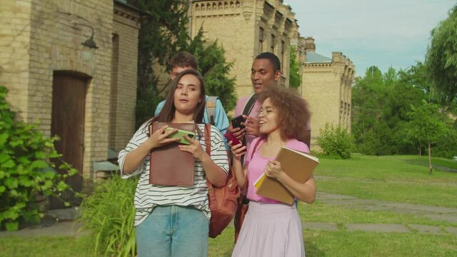 Close-up of cheerful mixed race students in casual clothing with copyboks walking along college building, browsing mobile app via internet and photographing together outdors at sunset