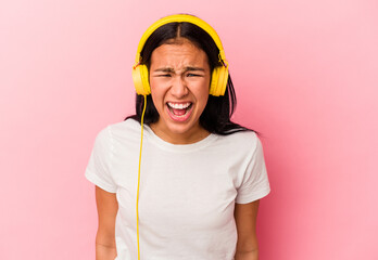 Young Venezuelan woman listening to music isolated on pink background screaming very angry and aggressive.