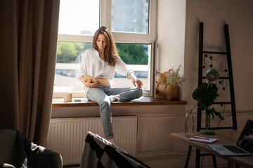 Young beautiful woman student reading book at home while sitting on window in modern apartment