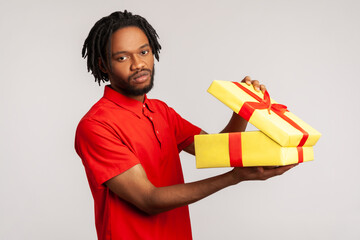 Disappointed man with dreadlocks in red casual T-shirt, holding unpacked gift box, has frustrated dissatisfied facial expression, upset with present. Indoor studio shot isolated on gray background. - obrazy, fototapety, plakaty