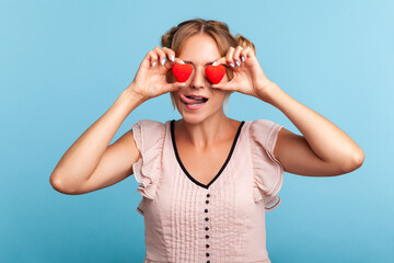 Loving eyes. Beautiful young adult woman with two funny hair buns holding two valentine hearts in front of her eyes like glasses, shows tongue out. Indoor studio shot isolated on blue background.