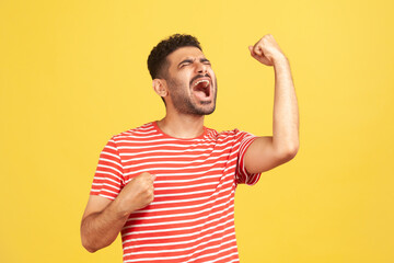 Fototapeta premium Extremely excited overjoyed man with beard in striped t-shirt shouting making yes gesture, amazed with his victory, triumph. Indoor studio shot isolated on yellow background